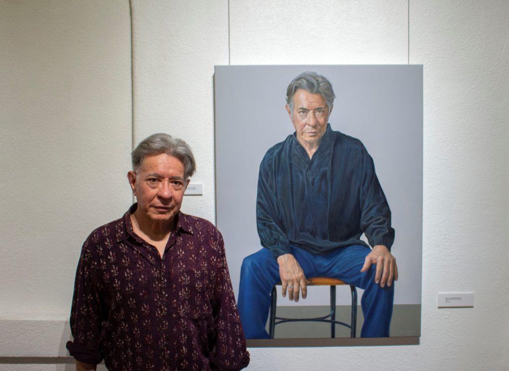 Artist Eloy Torrez stands next to his self-portrait among 19 other painted portraits that include his wife, late father, and friends. Photo credit: Rachel Cogswell