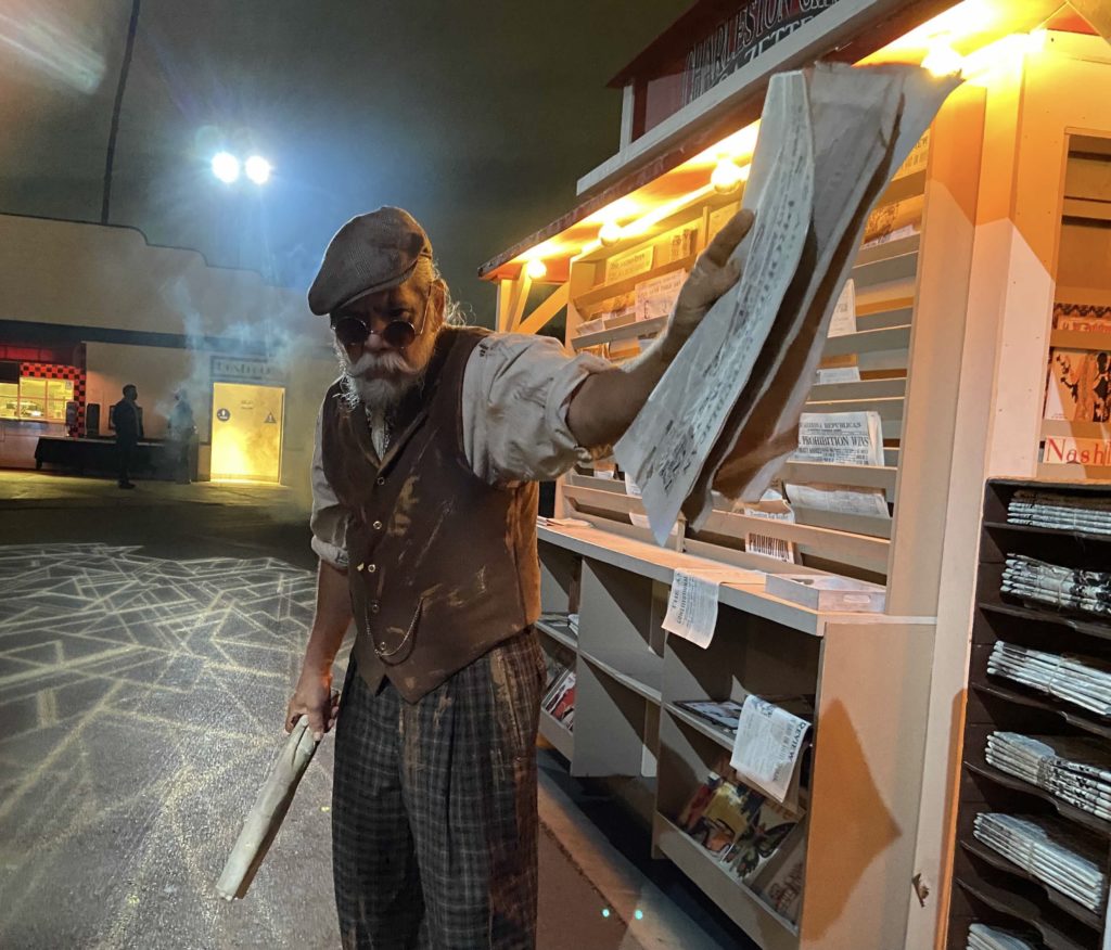 IMAGE, man with mustache and newspapers standing outside of a news stand. CAPTION, Gore-ing 20’s scare zone Newspaper Stand at Knott’s Scary Farm Photo credit: Dustin Malek