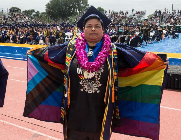 Erin Lacorte, at the 2022 Fullerton College Commencement. Erin wears sashes and Honors Cords. One is the Umoja Scholar sash (for Math studies); another is for Phi Theta Kappa, with Gold Tassel and Double Honors Cords for Lifetime member; an Associated Students stole; a royal-blue sash with FC monogram for Students of Distinction; a transfer medallion; the colorful linings to their robe is the Progress Pride Flag Photo credit: Fullerton College