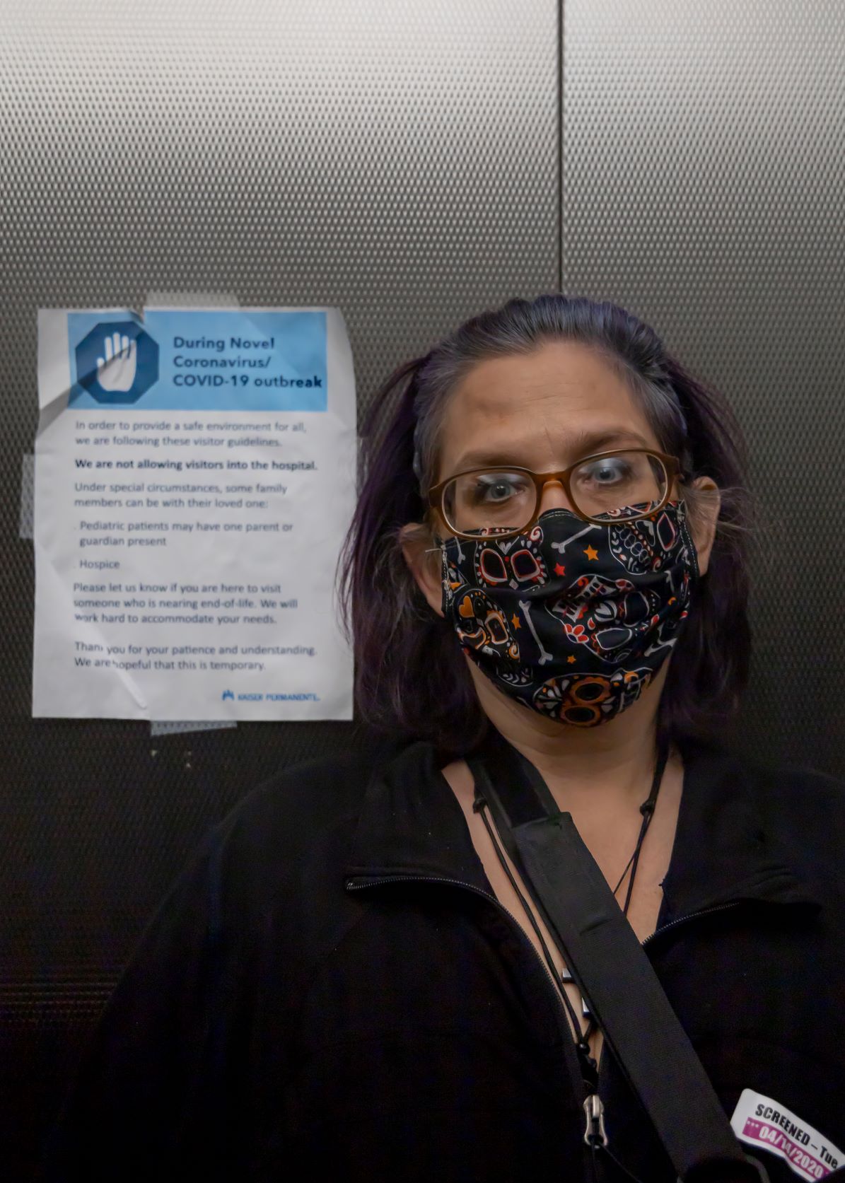 Anne Marie Flores, a visitor wears a mask and gloves standing in a Kaiser hospital elevator in front of a sign stating the rules regarding visitors during the COVID-19 pandemic.