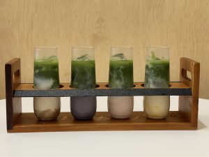 A Fruit Matcha Flight featuring banana, blueberry, guava, and mango flavors is served at Vivot Craft Cafe in Downtown Fullerton, Calif. The four-drink set is presented in a wooden tray.